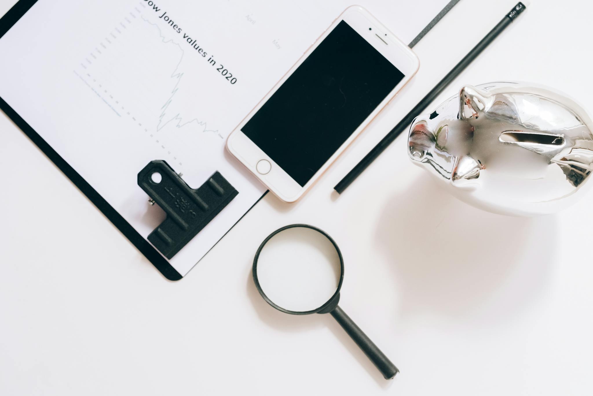 A flat lay of financial documents with a smartphone, magnifying glass, and piggy bank.
