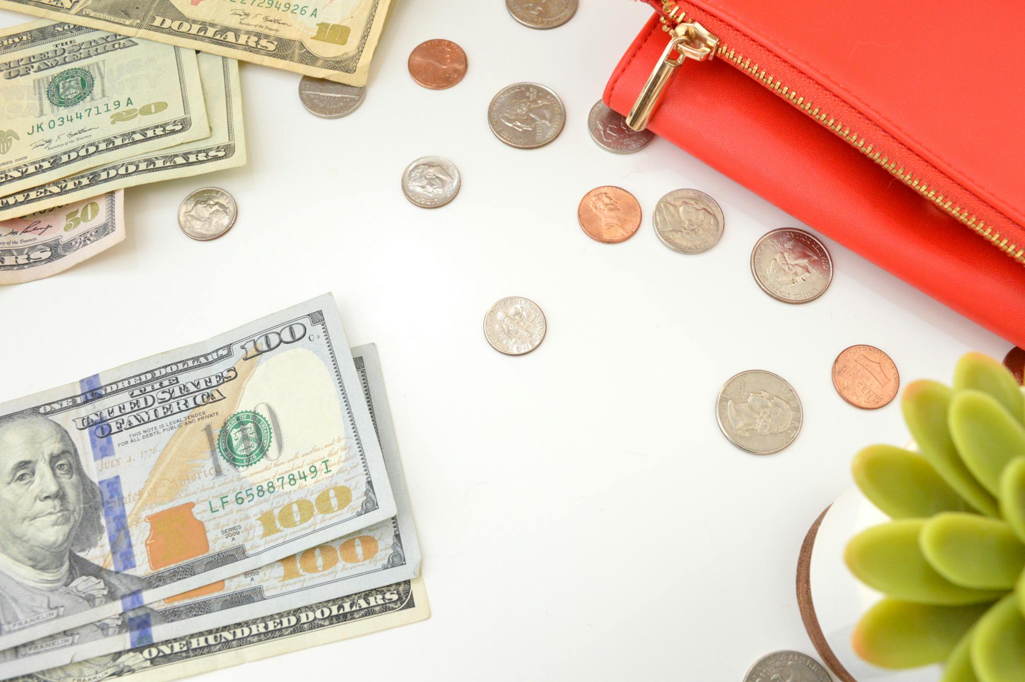 A top view of US dollar bills and coins scattered with a red wallet and succulent plant.